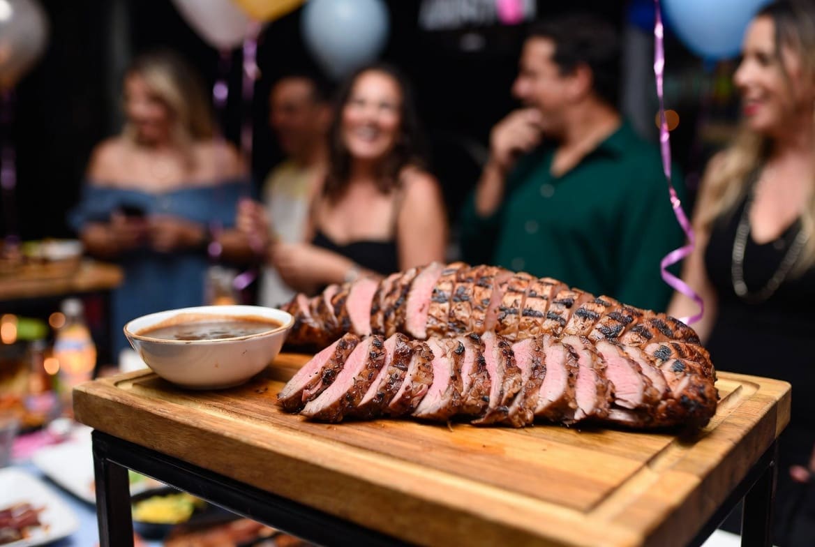 Sliced roasted meat from a spit braai served with gravy at a birthday party, blurred happy guests in background, professional celebration presentation