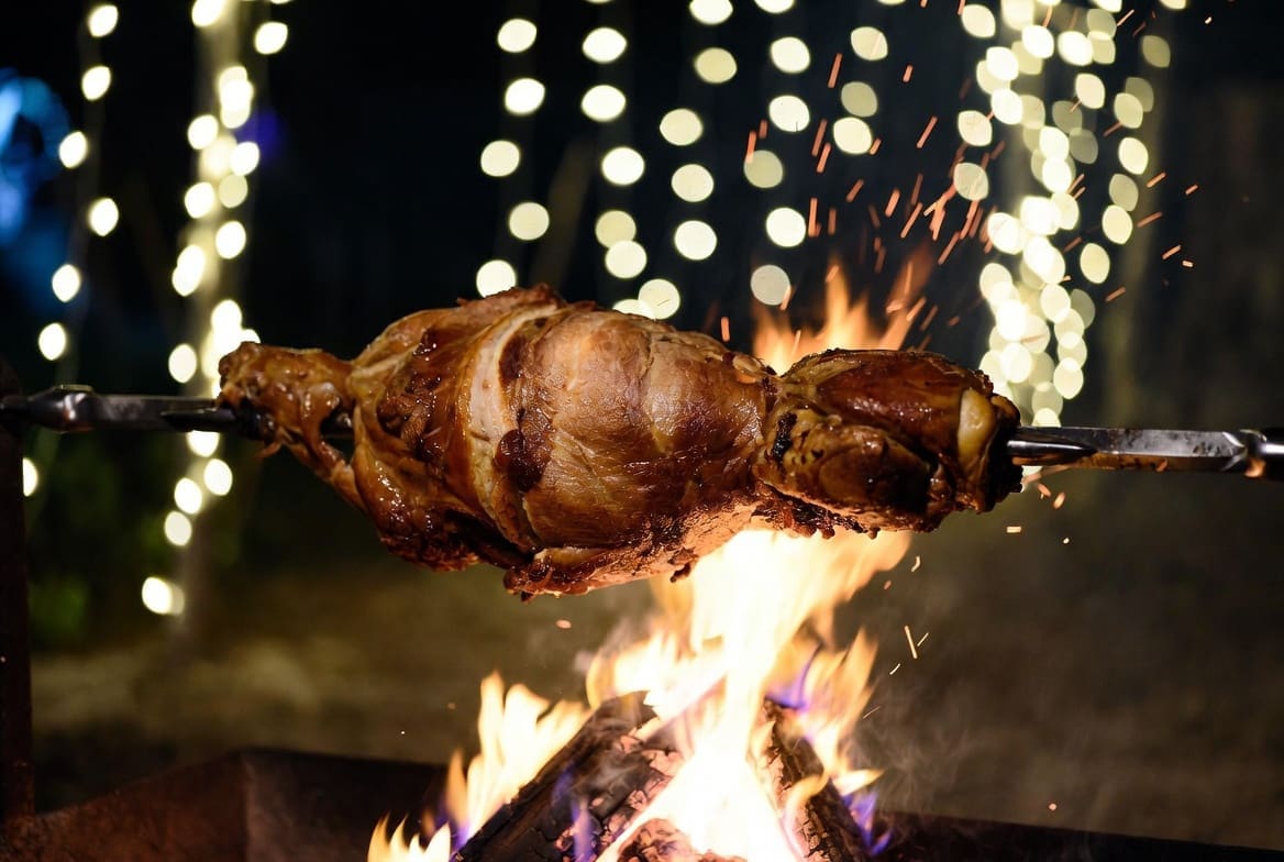 Roasted lamb on a spit braai South Africa at night with warm string lights and fire glow, cozy winter evening atmosphere, professional catering