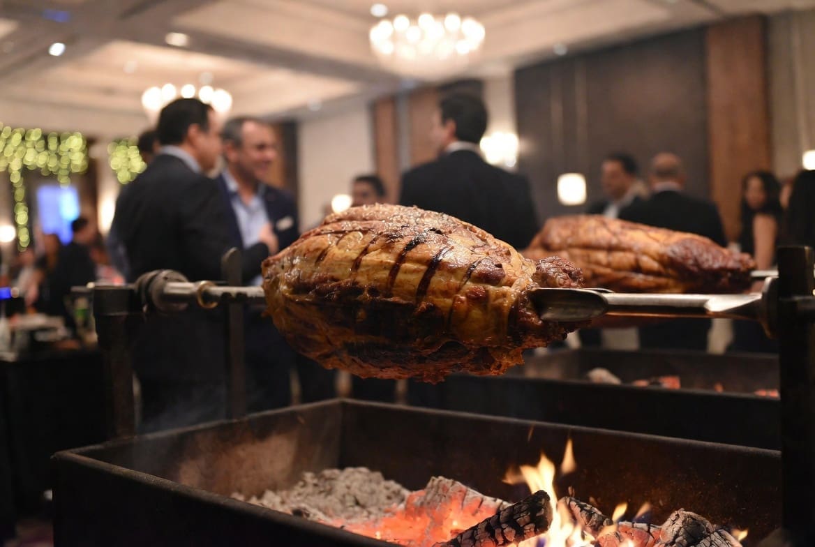 Roasted meat on a spit braai at an upscale corporate event in Johannesburg, blurred business people in background, food is the hero, premium catering