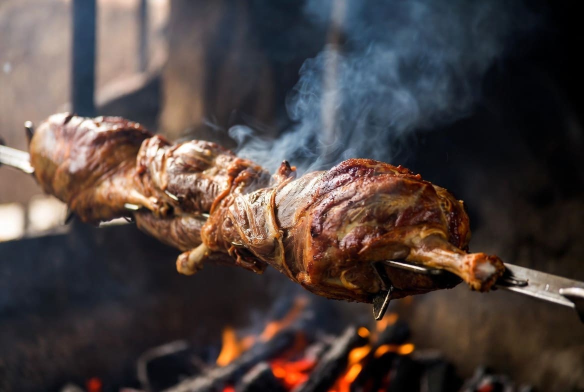 Crispy roasted lamb on a spit braai South Africa over glowing coals at night, steam rising, warm cozy winter atmosphere, professional catering