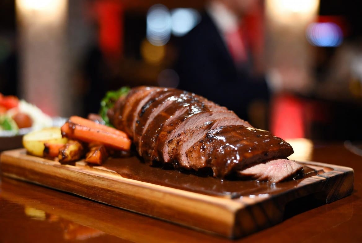 Sliced spit braai catering meat on a wooden board with rich gravy and roasted vegetables, professional presentation for events