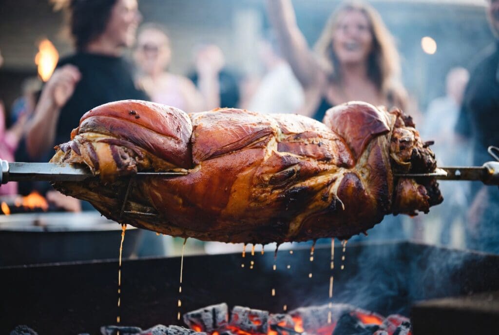 Whole roasted meat on a spit braai catering setup at an outdoor event in South Africa with blurred happy guests, premium professional presentation