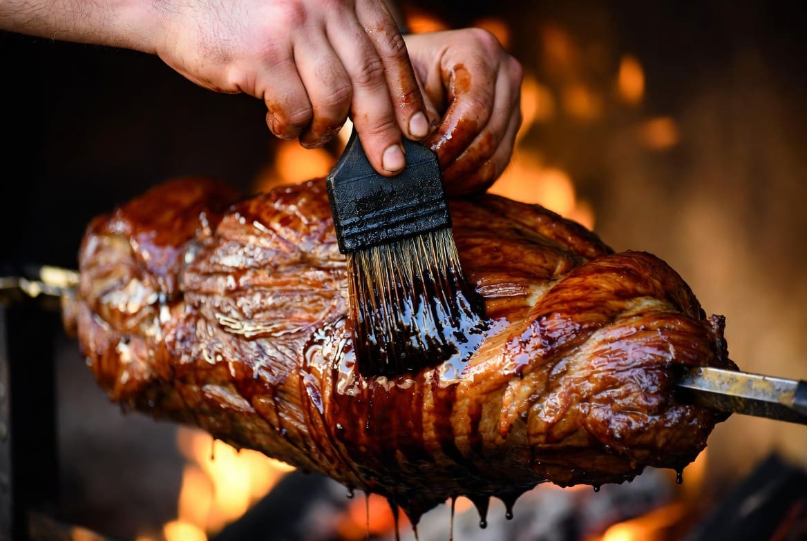 Close-up of a chef’s hands basting roasted spit braai meat with a quality brush, showing professional attention to detail