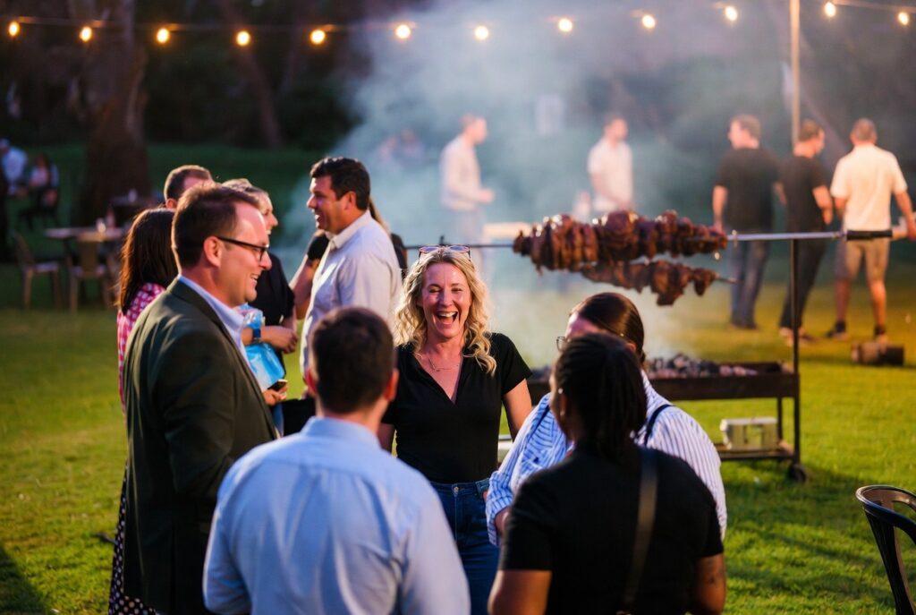 Guests socialising at a local outdoor event with a spit braai roasting in the background, warm evening atmosphere
