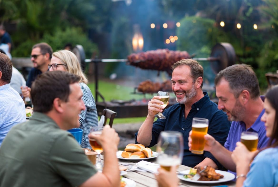Guests enjoying a meal at a long table during a local event, with a spit braai roasting in the soft-focus background