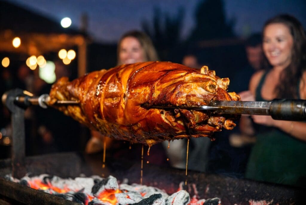 Professional spit braai packages featuring a whole roasted pork on the spit at an outdoor event with blurred guests, premium catering presentation